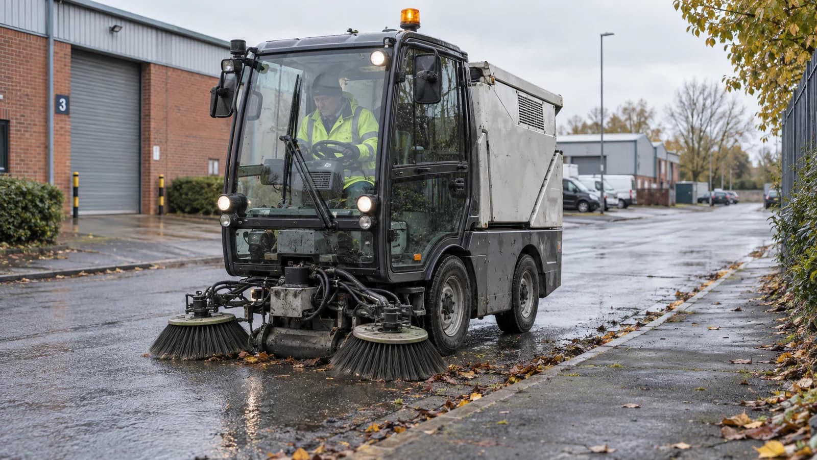 Compact road sweeper and operator cleaning a wet road on a UK industrial estate