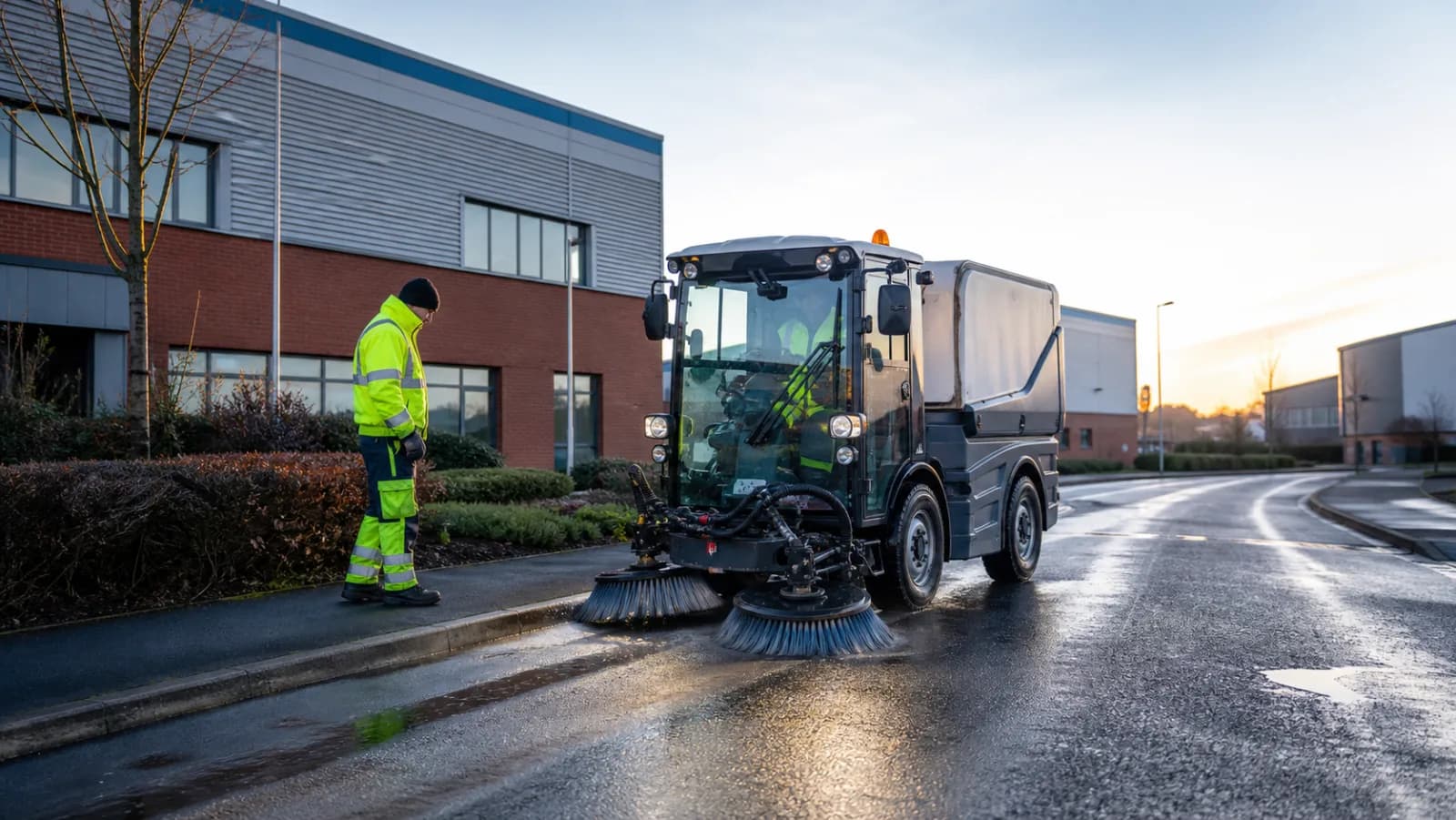 Road sweeper and high-visibility operator working on a wet UK industrial estate road